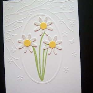 May include: A white card with a white embossed design of a tree branch with small white flowers and three white daisies with yellow centers in an oval frame.