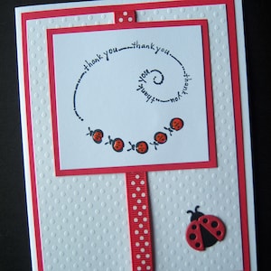 May include: A white card with a red border and red polka dot ribbon. The card has a red and black ladybug on the bottom and a black and white spiral design with the words "thank you" repeated around it. There are five red and black ladybugs on the spiral.