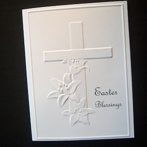 May include: White Easter greeting card with an embossed cross and lily flowers. The words "Easter Blessings" are printed in black script. The card has a rectangular shape with a raised border.