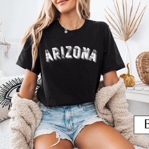 May include: Black t-shirt with the word "ARIZONA" in a distressed, white, and gray font. The model is wearing denim shorts and a cream-colored sweater. The image also includes decorative pumpkins and a pillow.