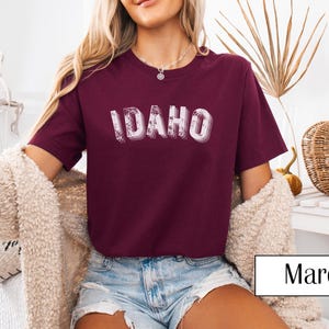May include: A maroon t-shirt featuring the word "IDAHO" in a layered, distressed font. The shirt is paired with denim shorts. The image is well-lit, with a neutral background. The word "Maroon" is in a white rectangle.