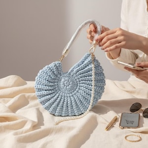 May include: A light blue, handmade crochet handbag with a circular design. The bag features a white braided handle and a pearl-beaded strap. Gold-colored hardware accents the bag. The bag is displayed on a cream-colored fabric.