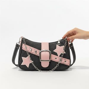 Puede incluir: Un bolso de hombro de mezclilla negra con detalles en rosa. El bolso presenta una correa rosa con hebilla plateada, dijes en forma de estrella rosa y una cadena plateada. El bolso tiene una correa negra con costuras blancas.