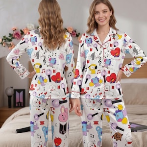 Pode incluir: Conjunto de pijama branco com um padrão colorido com personagens de desenhos animados, notas musicais e microfones. A parte de cima de manga comprida tem acabamentos e botões vermelhos. As calças a condizer completam o conjunto. O pijama é feito de material sedoso.