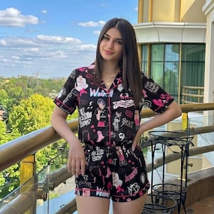 May include: Black pajama set with a pink trim and a pattern of cartoon graphics and text. The short-sleeved top and shorts feature a button-down front and drawstring waist. The word "Weird" is visible on the top.
