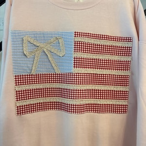 Könnte beinhalten: Ein hellrosa Sweatshirt mit einem Patchwork-Design, das einer amerikanischen Flagge ähnelt. Das Flaggen-Design umfasst blau-weiß gestreiften Stoff mit einer Spitzenschleife und rot-weißem Vichy-Stoff mit Spitzenbesatz.