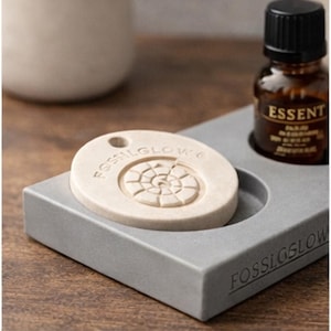 Può includere: Un vassoio grigio con la scritta "FOSSILGLOW" contiene un diffusore beige a forma ovale con un motivo a spirale e una piccola bottiglia di olio essenziale. La bottiglia ha un tappo nero e la parola "ESSENT" sull'etichetta.