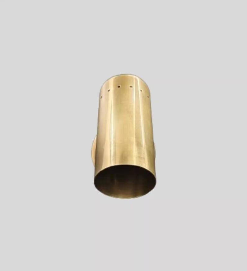 May include: A cylindrical, brass-colored wall sconce with a flared base. The sconce has a series of small holes near the top and a circular mounting plate on the side. The design is minimalist and modern.