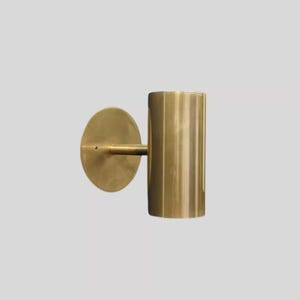 May include: A brass wall sconce with a cylindrical shade and a circular backplate. The sconce has a minimalist design and a warm, metallic finish. The fixture is mounted on a light gray wall.