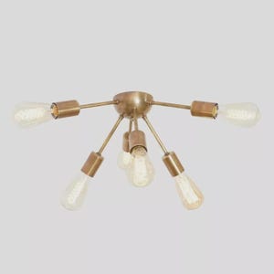 May include: A brass ceiling light fitting featuring six Edison-style bulbs. The fixture has a central hub with arms extending outwards, each holding a bulb. The clear bulbs have visible filaments, and the fitting is set against a light background.
