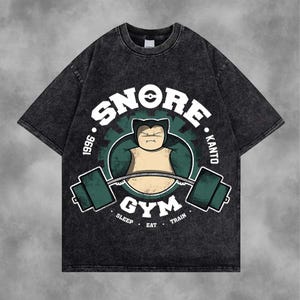Può includere: T-shirt grigio scuro dall'aspetto invecchiato. La parte anteriore presenta una grafica di un personaggio dei cartoni animati che solleva pesi, con le parole "SNORE GYM" e "SLEEP, EAT, TRAIN" in un design circolare. È incluso anche il testo "1996 KANTO".