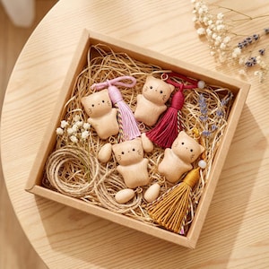 May include: A square gift box containing wooden cat keyrings with colourful tassels. The keyrings are light brown and have simple facial features. The box also contains twine, dried flowers, and straw.