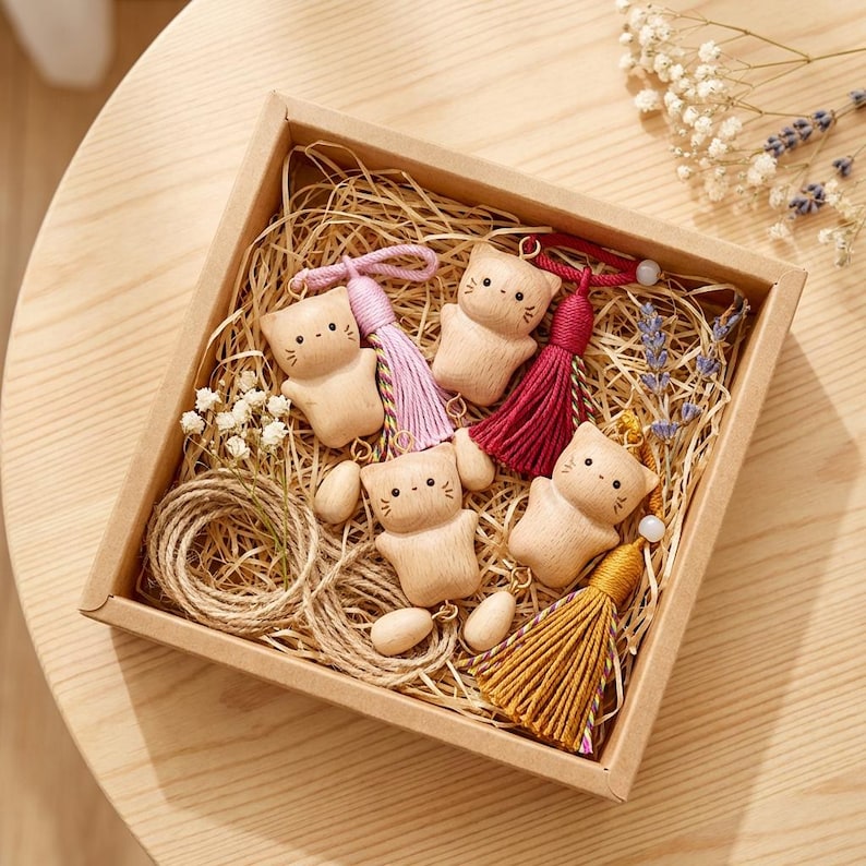 May include: A square gift box containing wooden cat keyrings with colourful tassels. The keyrings are light brown and have simple facial features. The box also contains twine, dried flowers, and straw.