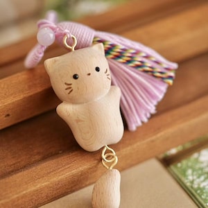 May include: A wooden cat charm with a light brown finish, featuring black eyes and whiskers. The charm is attached to a pink tassel with a multicolored accent and a gold-coloured metal ring. The charm is approximately 5 cm in height.
