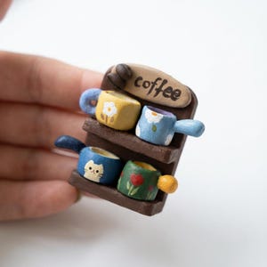 Pode incluir: Uma prateleira em miniatura com tema de café com quatro pequenas canecas coloridas. As canecas são pintadas com desenhos florais e de gatos. A prateleira é marrom com a palavra "coffee".