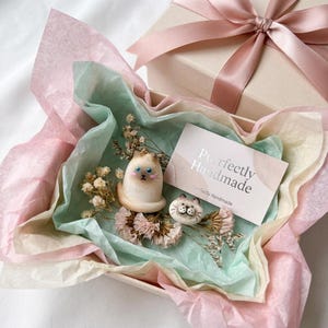Op de afbeelding: Een cadeauverpakking met een roze lint bevat twee kattenbeeldjes, droogbloemen en een kaart met de tekst "Purrfectly Handmade". De beeldjes zijn crèmekleurig met blauwe ogen en bruine aftekeningen.