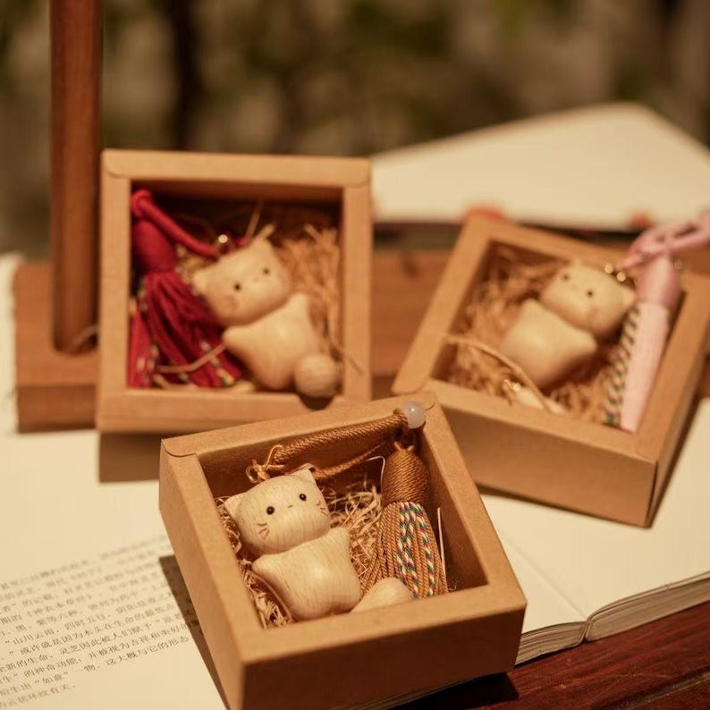 May include: Three small, square, cardboard boxes each contain a wooden cat figurine, decorative tassels, and straw-like filler. The cat figurines are light-coloured with simple facial features. The tassels are red, pink, and brown.