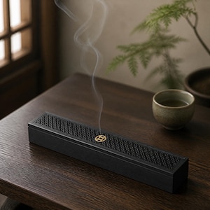 Puede incluir: Una caja quemadora de incienso rectangular de madera negra con un intrincado patrón de celosía en la tapa y un pequeño emblema circular de color dorado en el centro. Un fino rastro de humo blanco se eleva verticalmente desde el centro.
