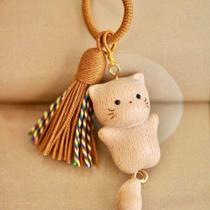May include: A wooden cat charm with black eyes and whiskers, suspended from a gold-coloured tassel and cord. The tassel has gold, green, blue, and purple threads. The charm is made of light-coloured wood.