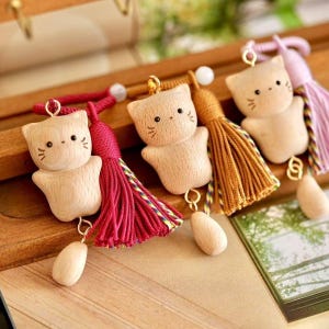May include: Wooden cat-shaped keyrings with detailed facial features and attached tassels in red, gold, and purple. Each keyring has a small wooden teardrop charm. The keyrings are displayed on a wooden surface.