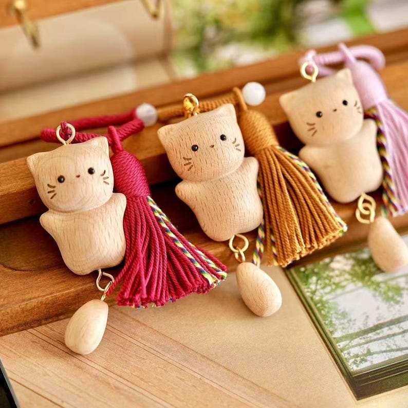 May include: Wooden cat-shaped keyrings with detailed facial features and attached tassels in red, gold, and purple. Each keyring has a small wooden teardrop charm. The keyrings are displayed on a wooden surface.