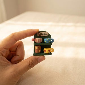 Pode incluir: Miniatura de exposição de café verde com duas prateleiras. A prateleira superior contém uma caneca listrada e uma caneca azul com bolinhas. A prateleira inferior tem uma caneca floral e uma caneca amarela em forma de coração. A palavra "Coffee" está escrita acima da prateleira superior.