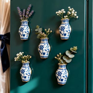 Könnte beinhalten: Fünf kleine Vasen aus blau-weißem Porzellan mit floralen Mustern, jede mit verschiedenen Blumen und Grünpflanzen. Die Vasen sind auf einer dunkelgrünen Oberfläche angebracht und bilden eine dekorative Präsentation.