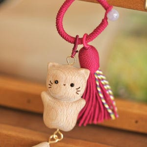 May include: A wooden cat charm with a pink tassel and cord. The cat has black eyes and whiskers. The charm is suspended from a pink cord with a small bead.