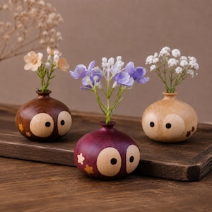 Op de afbeelding: Drie kleine, ronde houten vaasjes in de vorm van uilen, elk met grote ronde ogen en kleine steraccenten. De vaasjes zijn donkerbruin, paars en lichtbeige en bevatten elk een klein boeket kunstbloemen in perzik, paars en wit.