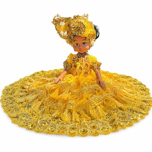 May include: A doll with dark hair wearing a bright yellow dress and matching headpiece. The dress features a wide, circular skirt adorned with gold sequins, intricate lace trim, and dangling fringe accents. The bodice is embellished with gold beads.