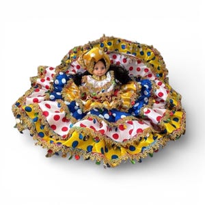 Op de afbeelding: Een pop in een kleurrijke jurk met een gele hoofdbedekking. De jurk heeft lagen van witte, blauwe en gele stof met rode en blauwe stippen. Gouden randen en decoratieve accenten maken de feestelijke look compleet.