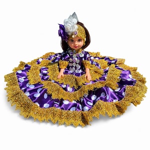 May include: A doll wearing a purple satin dress with white polka dots and gold metallic fringe trim. The doll features a matching gold headpiece with a white and purple flower, and a bodice decorated with clear, faceted, oval-shaped stones.