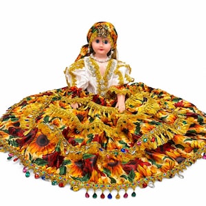 May include: A doll wearing a white blouse with gold trim and a wide, full skirt featuring a sunflower print. The doll has a matching headpiece, a beaded necklace, and the skirt is decorated with gold fringe and colorful hanging crystal beads.