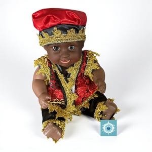 May include: A dark-skinned doll wearing a red and black hat with gold trim. The doll is dressed in a red vest with gold accents and black trousers, also with gold trim. The doll is sitting with its legs bent and arms outstretched.