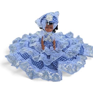 May include: A doll with dark skin and curly black hair wearing a blue and white gingham dress with multiple layers of white lace trim. The doll features a matching headpiece adorned with a silver-toned decorative brooch and a white pearl accent.