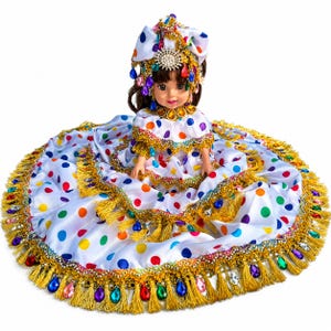 May include: A doll with brown hair wearing a white dress featuring multicolored polka dots, gold fringe trim, and dangling teardrop-shaped gems. The doll also wears a matching headpiece adorned with gold trim, colorful gems, and a central silver brooch.