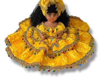 May include: A doll wearing a bright yellow dress with a ruffled skirt and a matching head covering. The dress is trimmed with gold and colorful beads. The doll has black curly hair and blue eyes.