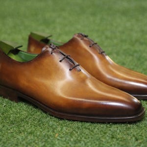 Puede incluir: Un par de zapatos de vestir de cuero marrón con cordones marrones oscuros. Los zapatos tienen un acabado pulido y están sobre una superficie verde. El interior del zapato es verde.