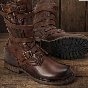 Könnte beinhalten: Ein Paar wadenhohe Stiefel aus braunem Leder mit Used-Optik, mehreren umlaufenden Riemen mit messingfarbenen Metallschnallen und einer dicken, dunklen Gummiprofilsohle. Das Leder weist durchgehend natürliche Farb- und Strukturvariationen auf.