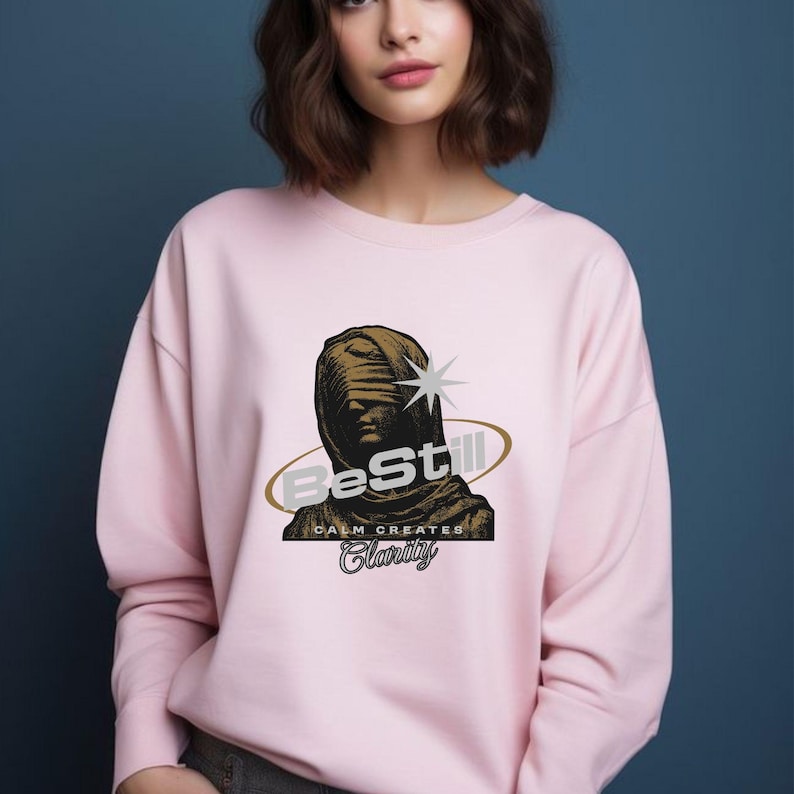 May include: A light pink sweatshirt featuring a graphic of a bust with the text "Be Still" and "Calm Creates Clarity". The graphic is in shades of brown and gold. The sweatshirt is a crew neck style.