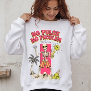 Op de afbeelding: Een wit sweatshirt met lange mouwen en een afbeelding van een skelet met stervormige zonnebril en gestreepte korte broek, met een drankje in de hand. De tekst luidt NO PULSE NO PROBLEM en life's better when you're dead inside, met een palmboom.