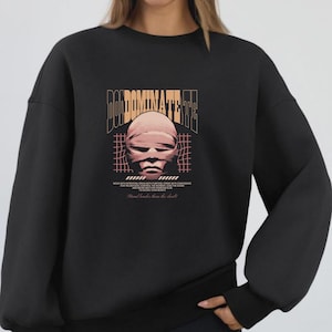 May include: Black sweatshirt with a graphic design featuring the text "DOMINATE THE" above a wrapped face illustration. Additional text at the bottom reads "Mind harder than the skull". The design is in shades of tan and pink.