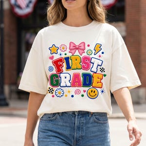 Op de afbeelding: Een crèmekleurig T-shirt met korte mouwen en een kleurrijke grafische print met de tekst FIRST GRADE in dikke, veelkleurige blokletters. Het ontwerp bevat een roze strik, een gele ster, een bliksemschicht, bloemen en een gele smiley.