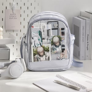 Peut inclure: Un sac à dos gris clair doté d'une poche frontale transparente remplie d'illustrations de personnages de style anime, d'accents de pellicule photo et de petites épinglettes décoratives. Le sac possède un compartiment principal zippé.