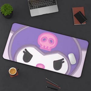 May include: A large desk mat with a cartoon character design in purple and white, featuring a pink skull and a stern expression. A computer mouse sits on the mat. Other items on the desk include a laptop, a notebook, and a cup of coffee.