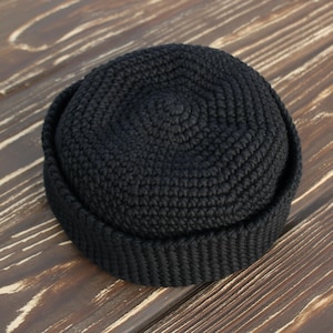 May include: A black, crocheted beanie hat with a rounded crown and a folded brim. The hat is made of a thick, textured yarn and sits on a wooden surface. The hat is a solid color and appears to be handmade.