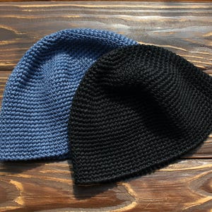 May include: Two crochet beanies, one in a solid denim blue colour and the other in a solid black colour. Both hats feature a consistent, dense stitch pattern throughout the rounded crown, providing a textured appearance and a simple, classic shape.