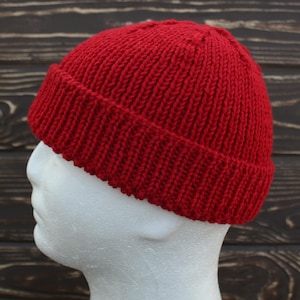 May include: A vibrant red knit beanie hat. The hat is made of a thick, textured yarn and features a folded brim. The hat is displayed on a white mannequin head against a dark wood background.