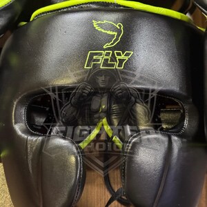 K&ouml;nnte beinhalten: Schwarzer Box-Kopfschutz mit neongr&uuml;nen Akzenten. Der Kopfschutz zeigt das Wort "FLY" in Neongr&uuml;n, eine Vogelgrafik und eine K&auml;mpferillustration. Dieser Kopfschutz dient dem Schutz beim Boxen und anderen Kampfsportarten.