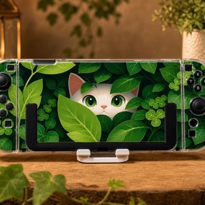 May include: A handheld gaming console featuring a clear protective case with a vibrant graphic print of a white cat peeking through lush green leaves and clover. The device includes black joysticks, directional buttons, and action buttons.